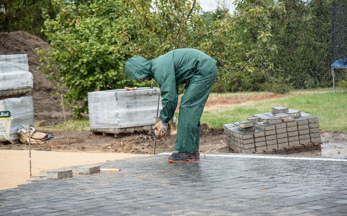 Pflasterer verlegt Betonsteine auf der Splittbettung, Paletten mit Steinen im Hintergrund